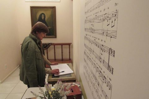 A irmã Helena Mary no hall de entrada do Memorial Santa Teresa Verzeri onde está o livro de visitas. Na parede, pauta da musica “Teresa Madre Nostra” cantada na Praça de 
São Pedro em 10 de junho de 2001 quando Teresa Verzeri foi canonizada pelo Papa João Paulo II (Obs. Não é possível realizar fotografias de outras salas do memorial)